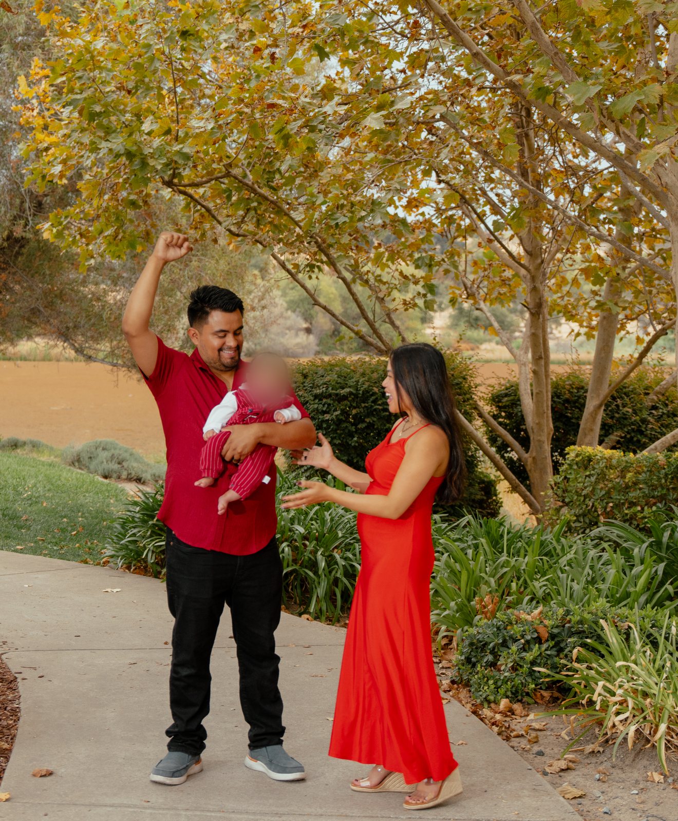 On a park path under yellowing trees, the father in a red shirt raises one fist with the baby in his other arm, while the mother in a long red dress laughs with her hands held out.