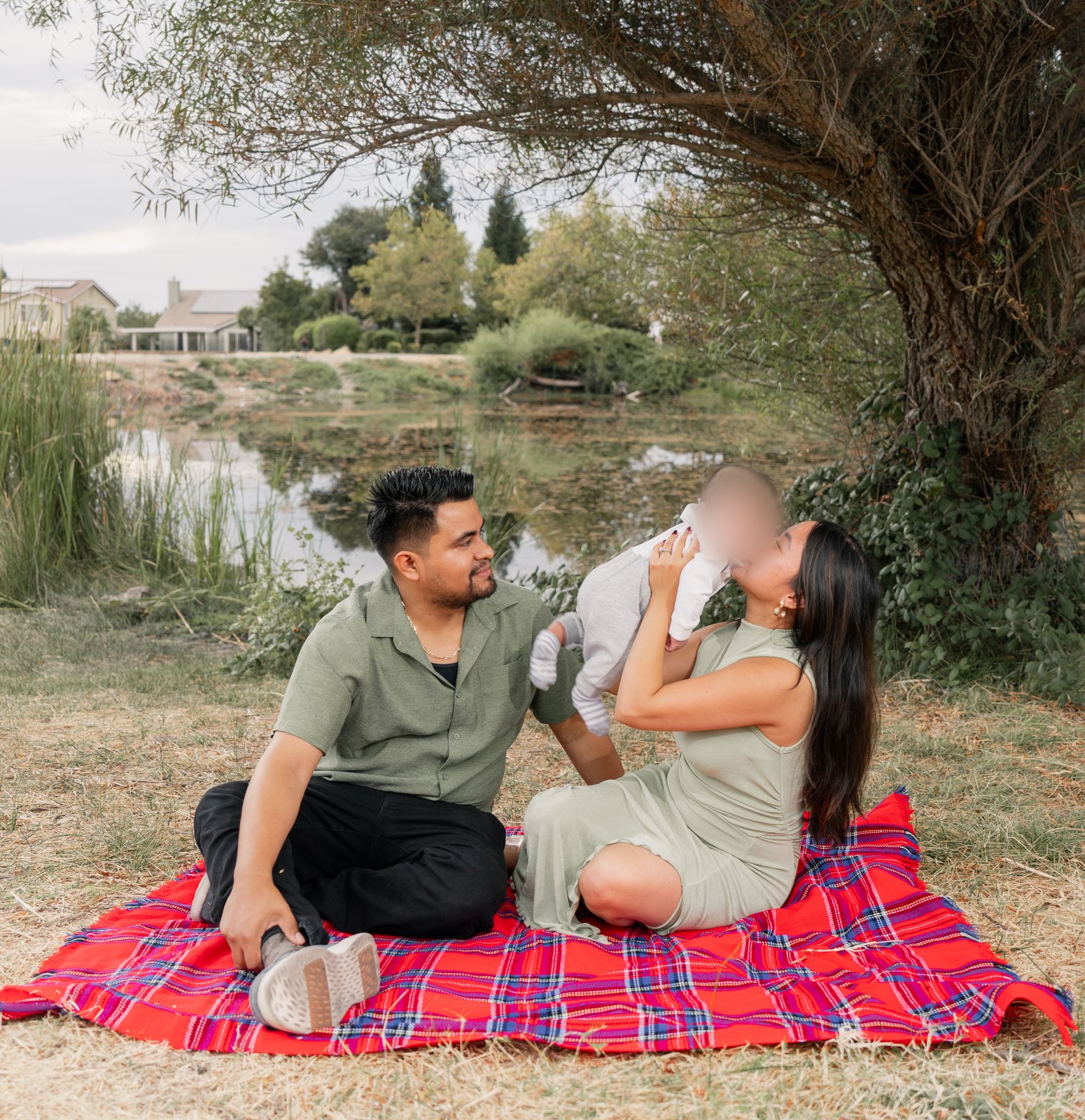The father and mother sit on a red plaid blanket under a large tree at the edge of a pond, and she lifts their baby up to her face for a kiss while he watches.