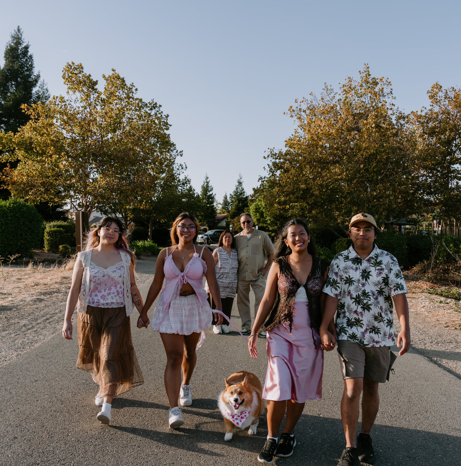 Four young adults walk toward the camera holding hands on a paved path lined with trees, a corgi in a pink bandana trotting between them on a leash, with the parents standing behind.