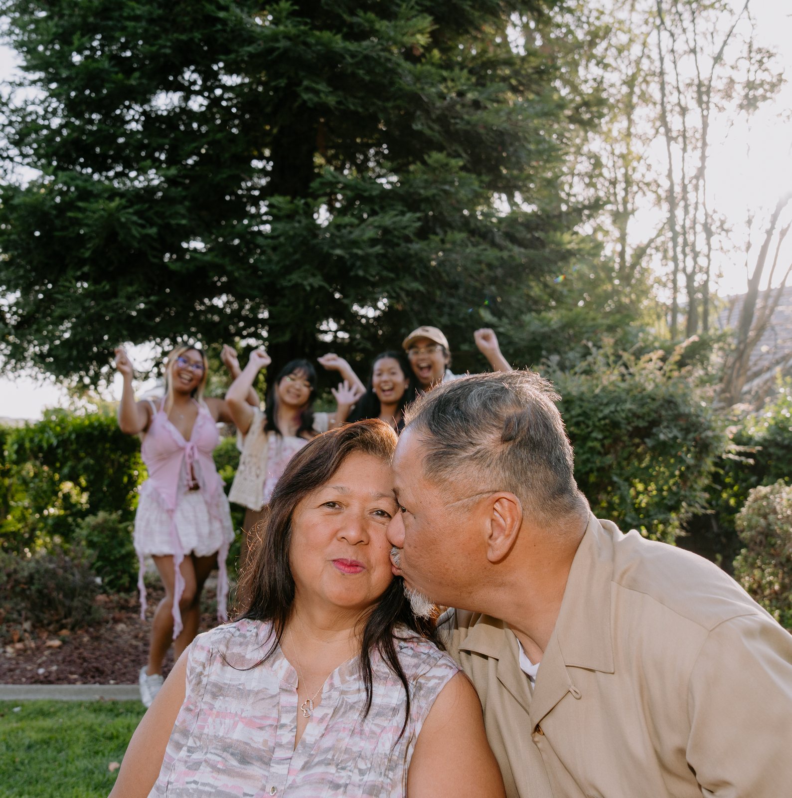 The father kisses the mother on the cheek in the foreground while four young adults behind them throw their arms in the air and laugh in front of a tall evergreen.