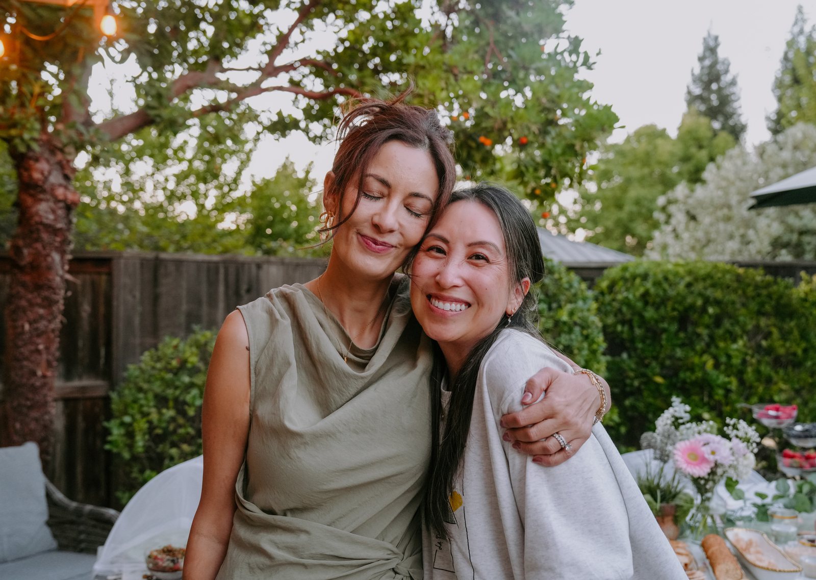 Two women stand cheek to cheek in the backyard, one in a sage green dress with her eyes closed and her arm around the other, who smiles at the camera.