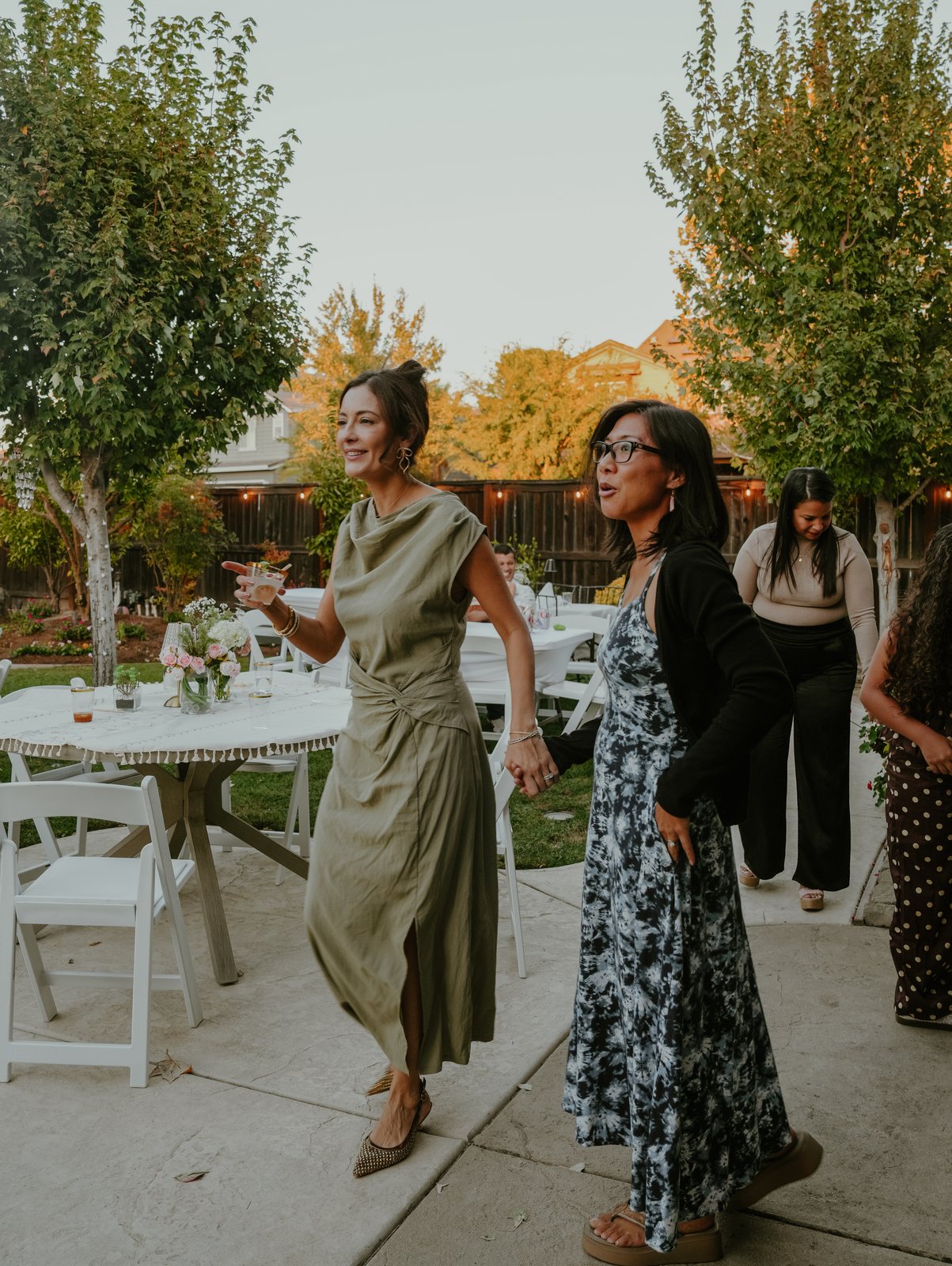 A woman in a sage green dress raises a glass as she steps across the patio hand in hand with a woman in a blue-and-white print dress.