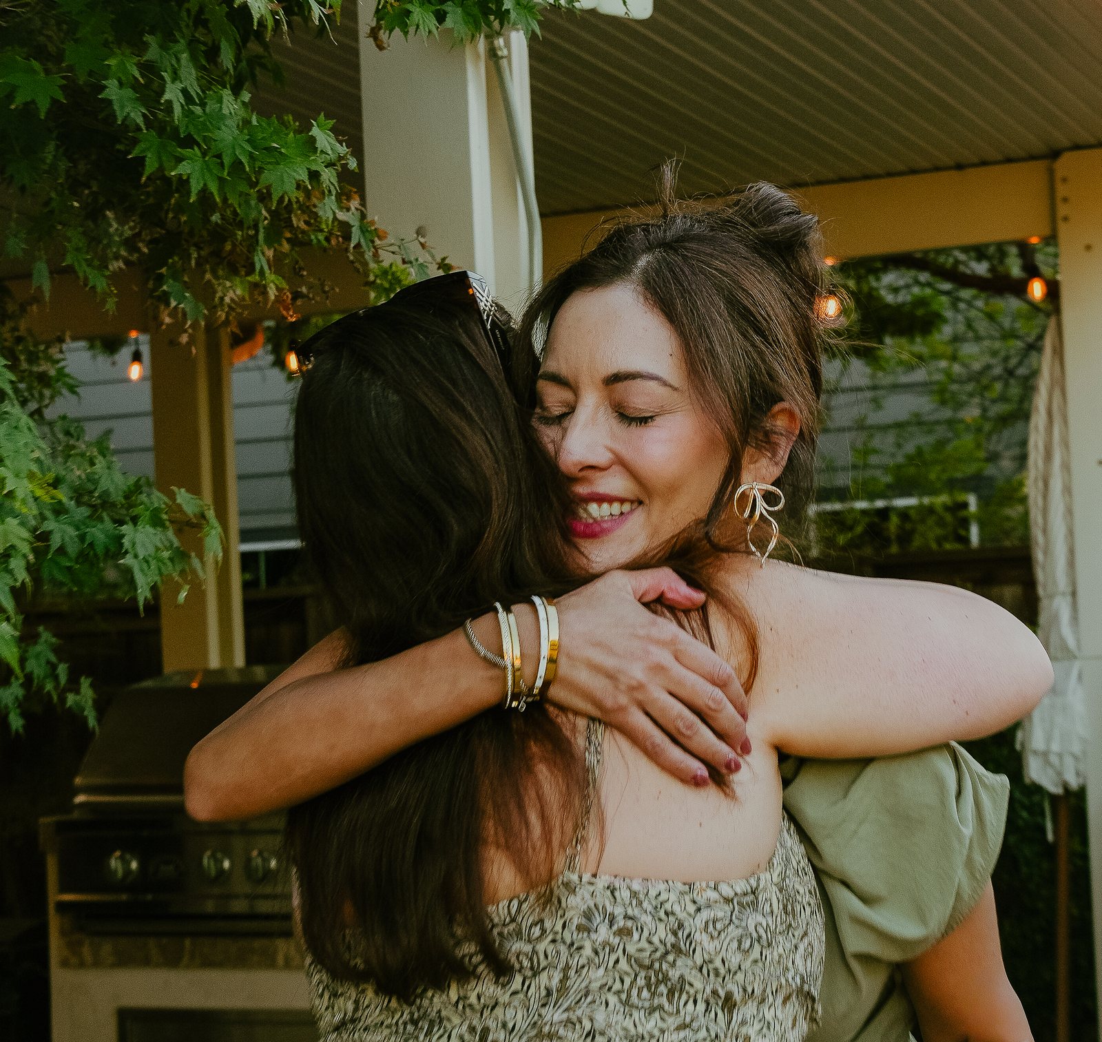 Two women hug under a covered patio strung with lights, one smiling with her eyes closed and gold bangles on the arm wrapped around the other woman.