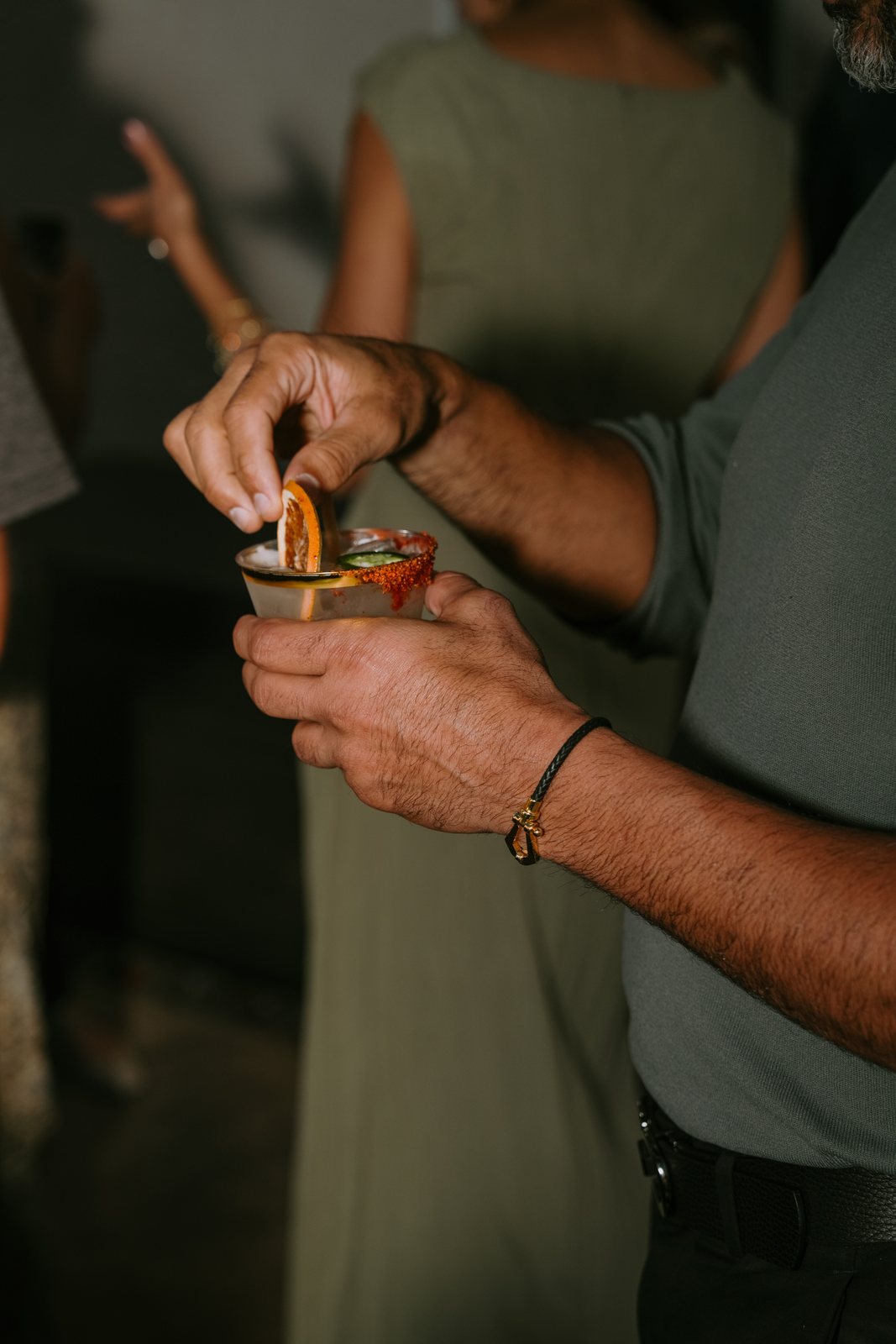 A man's hands set a citrus slice into a drink with a red rim, with a woman in a sage green dress behind him.