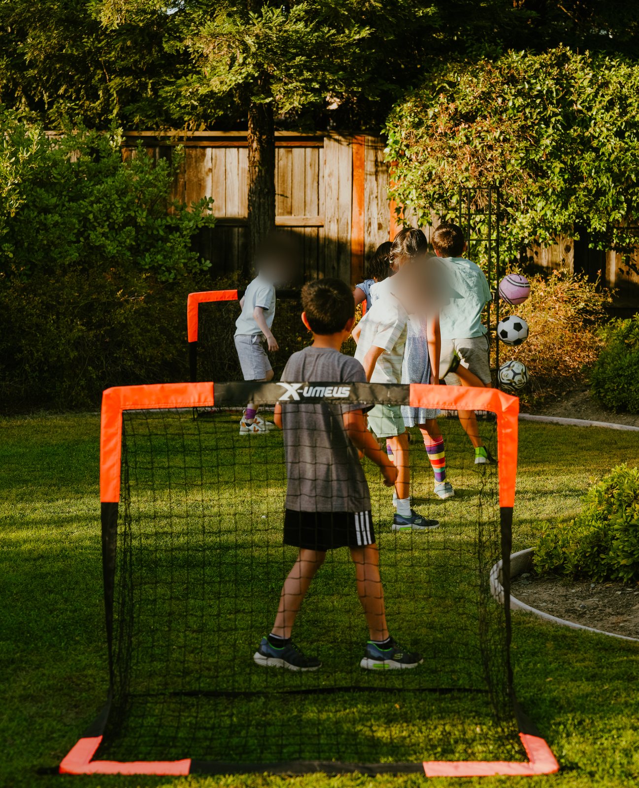 Seen from behind a small orange-framed soccer goal, a boy in a gray T-shirt stands in the net while other children gather by the fence across the lawn.