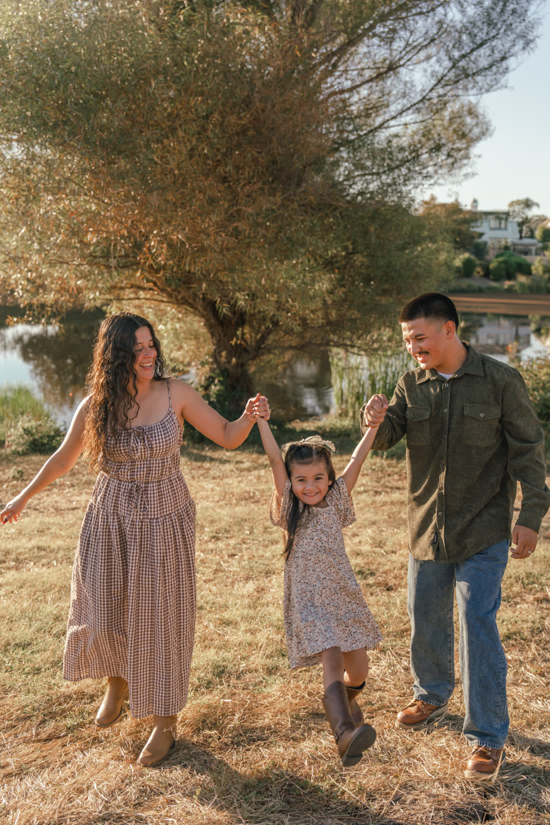 Laughing parents swing their daughter between them by the hands, her brown boots off the ground, in a dry grass field in front of a willow and a pond.