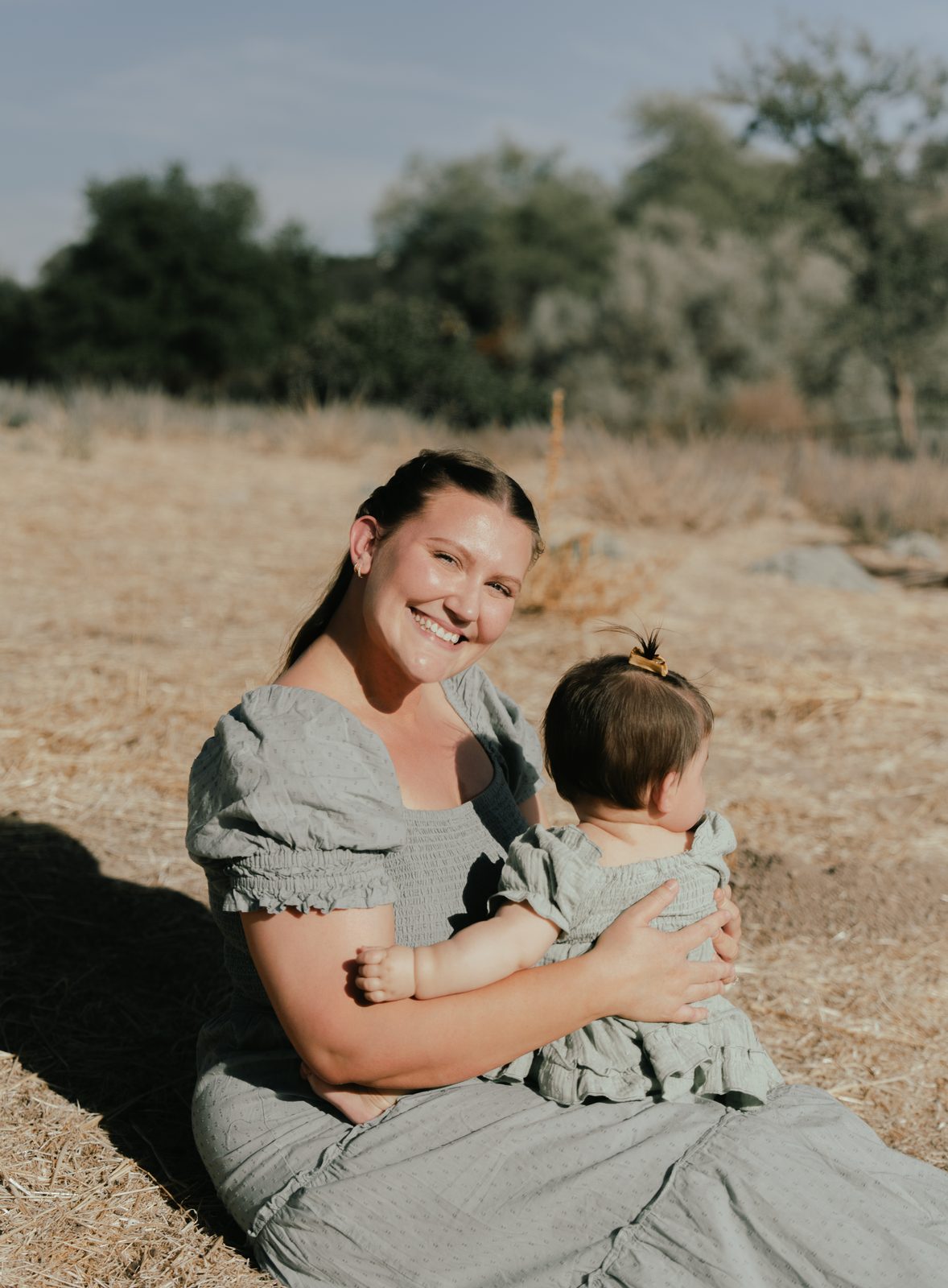 A mother sits in golden grass with her baby daughter on her lap, smiling at the camera