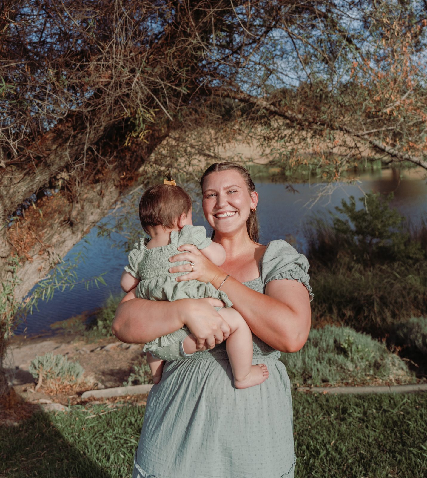 The mother stands under the arching branches of a tree with water behind her, smiling and holding the baby against her chest, the baby facing away with one bare foot showing.