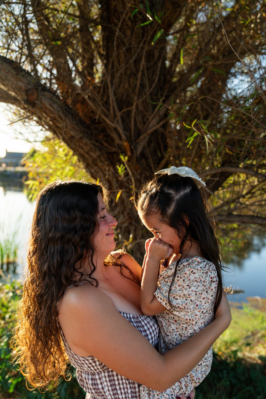 A mother in profile smiles at the daughter she is holding, who giggles with both fists pressed to her mouth, under a willow with the pond behind