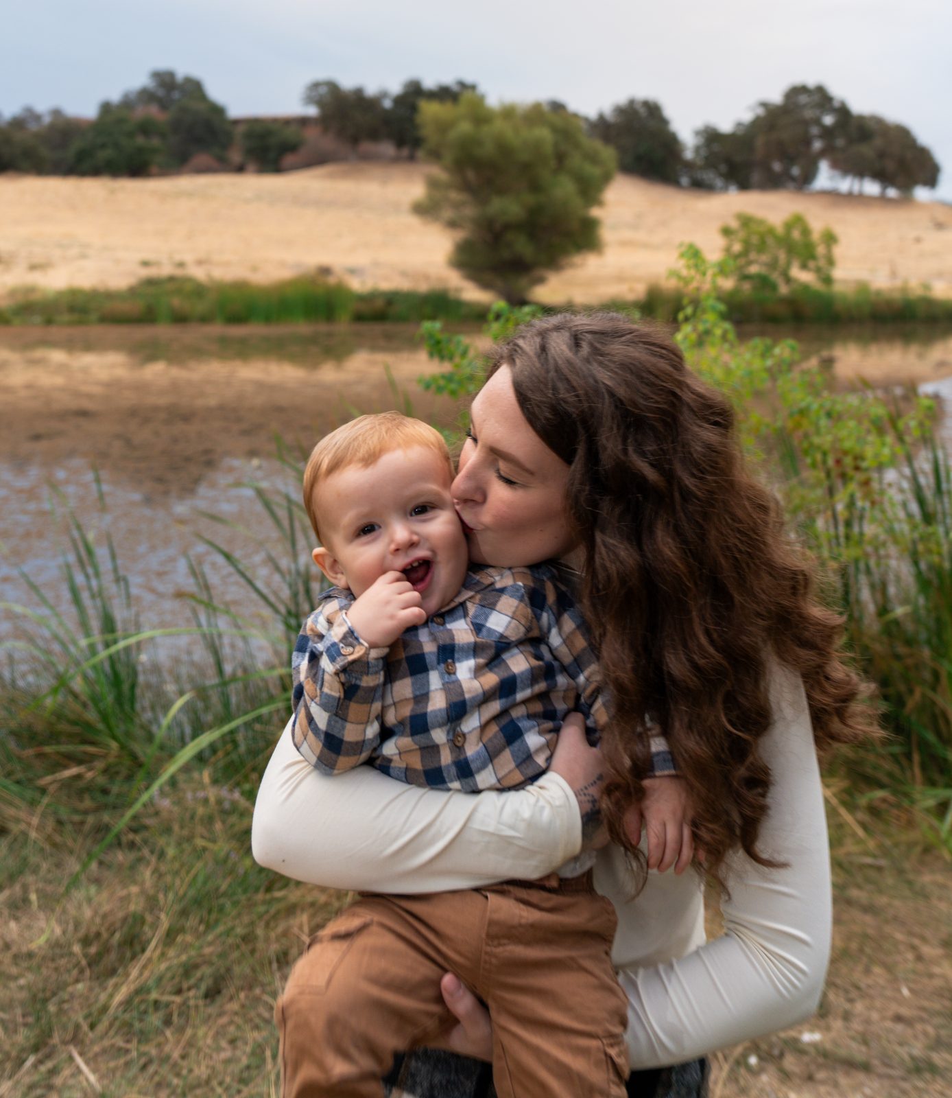 The mother, eyes closed, kisses the toddler's cheek as he laughs into the camera with a hand at his mouth, the pond and a dry hillside behind them.