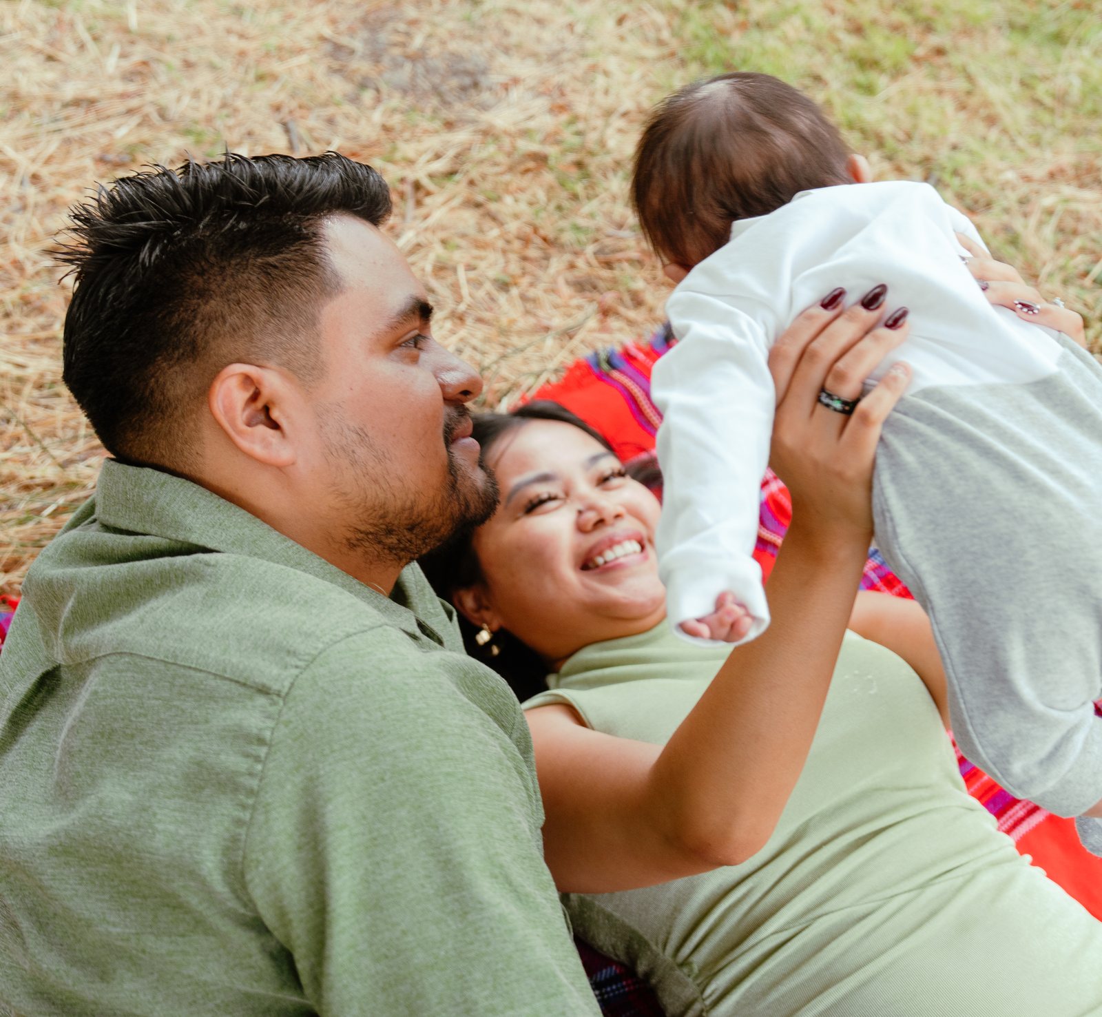 Lying back on the plaid blanket, the mother laughs as she holds their baby up above her, the back of the baby's head to the camera, with the father lying beside her.