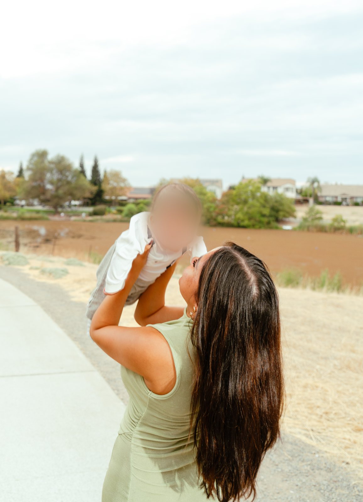 Seen from behind on a paved path, the mother lifts their baby high above her face, with bare brown ground, trees and houses beyond under a cloudy sky.