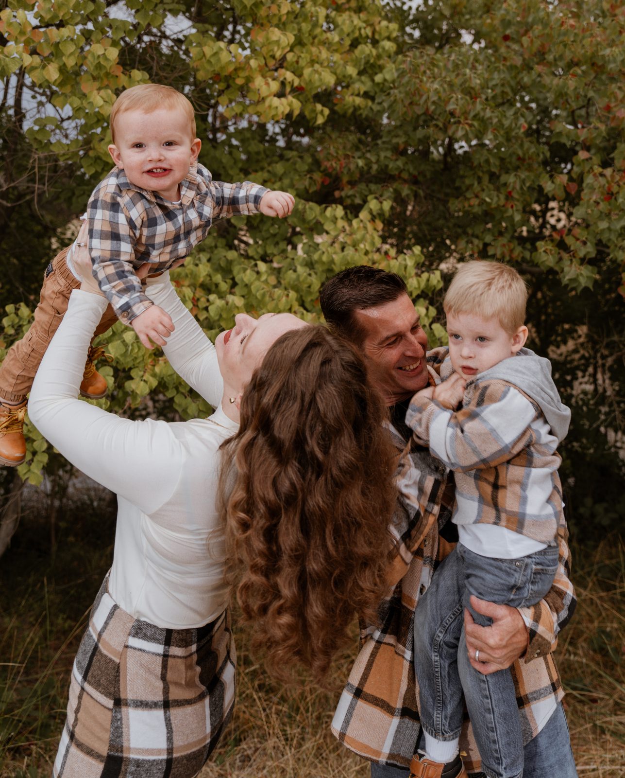 A mother tips her head back and lifts her toddler high overhead while the father, laughing, holds their older boy