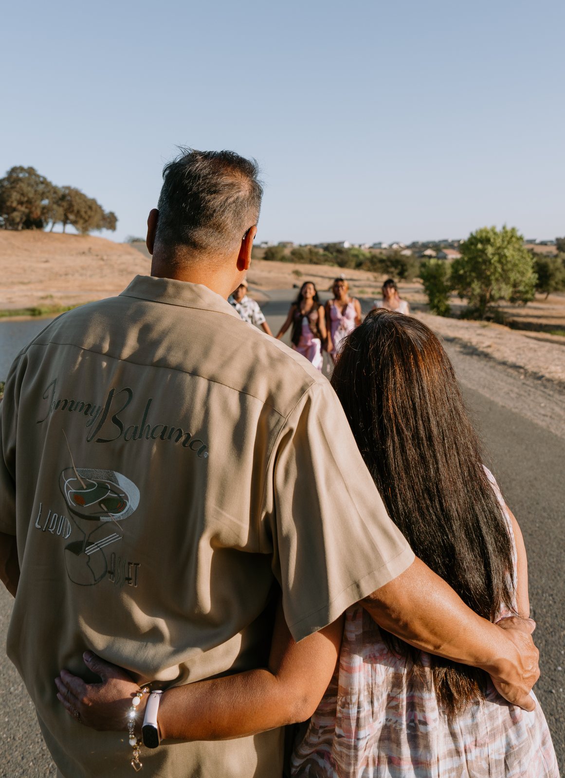 Seen from behind, the parents stand with their arms around each other on a path beside a pond, watching four young adults walk toward them past dry hills.