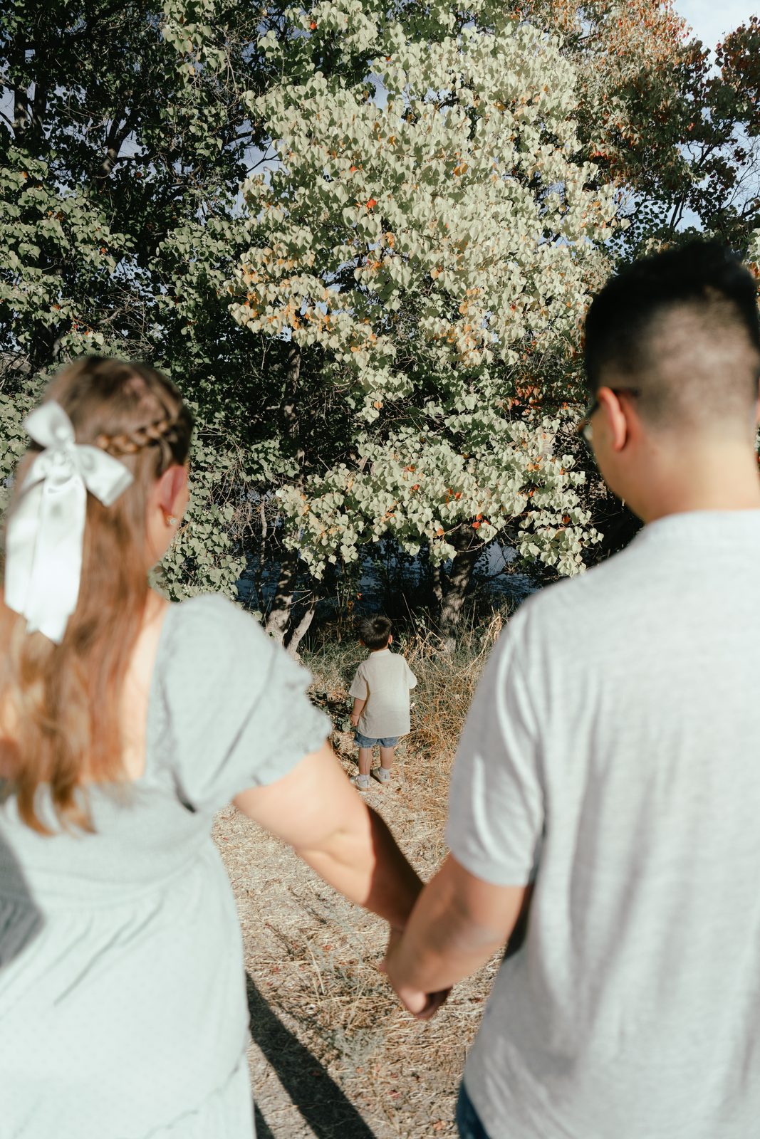 Between the out-of-focus shoulders of the parents holding hands, the toddler stands alone on a dry grass path in front of a large pale-leaved tree.