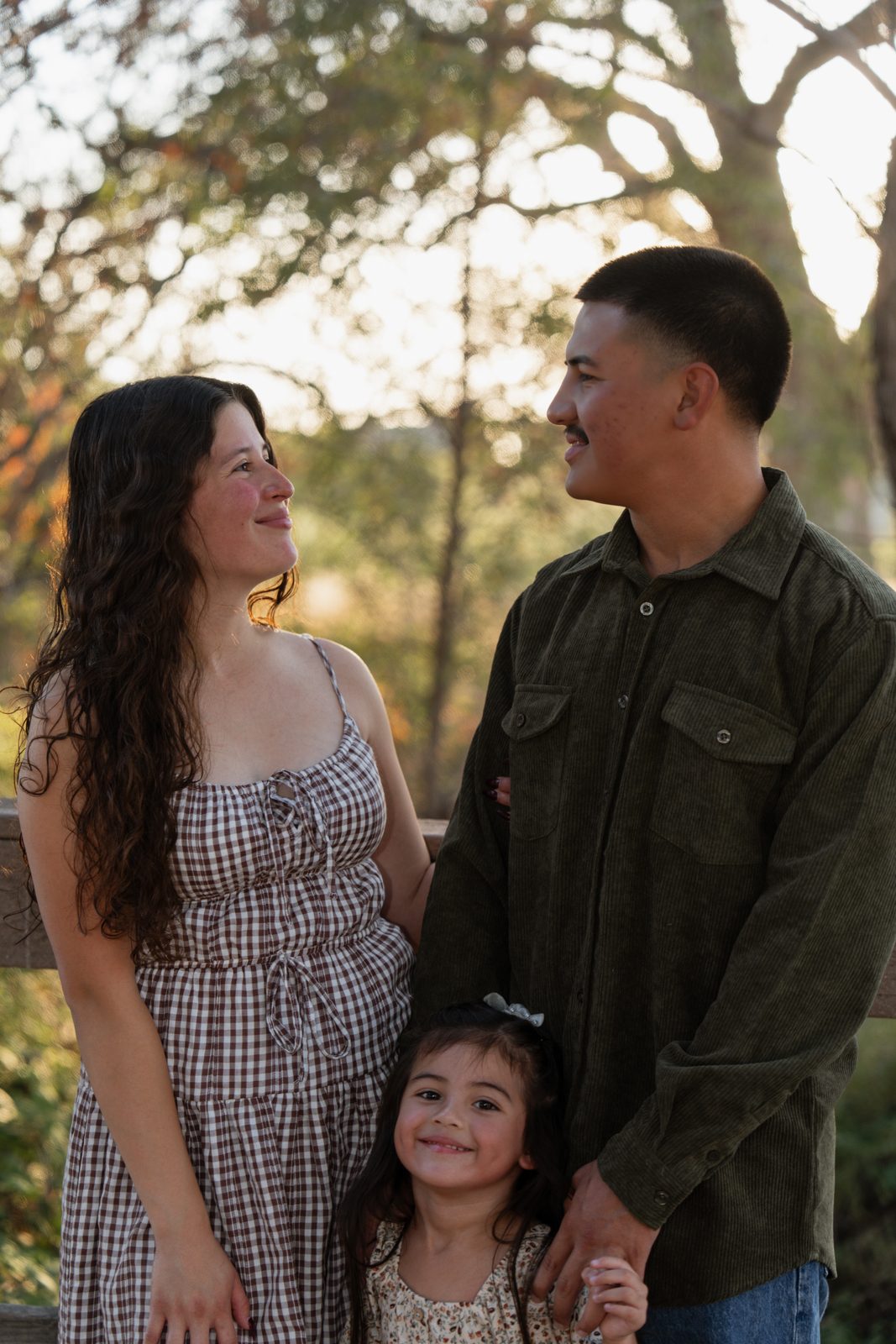 A mother and father smile at each other while their daughter, standing between them, grins up at the camera