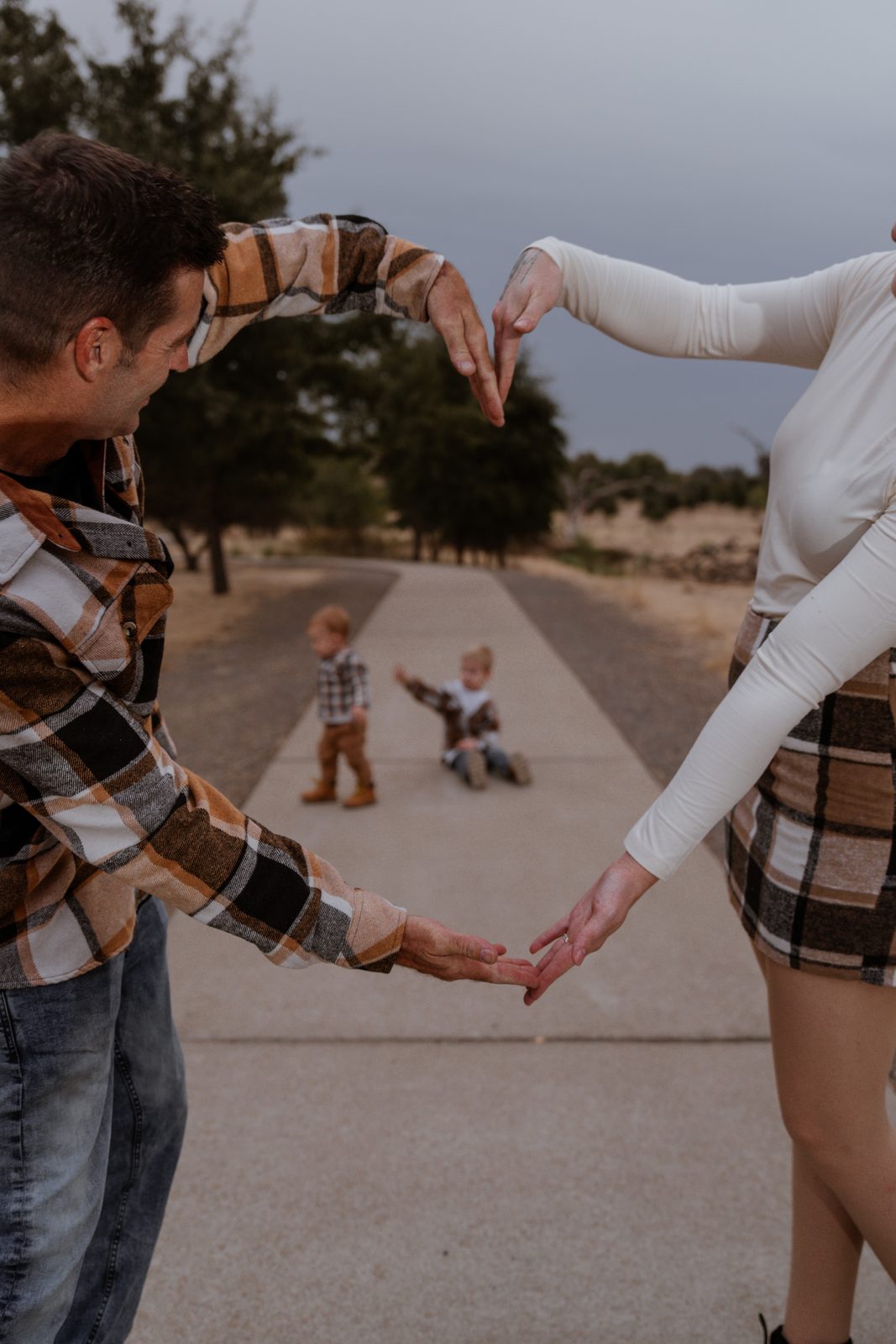 The parents' arms form a heart in the foreground, framing the toddler standing and the older boy sitting on a concrete path behind them.