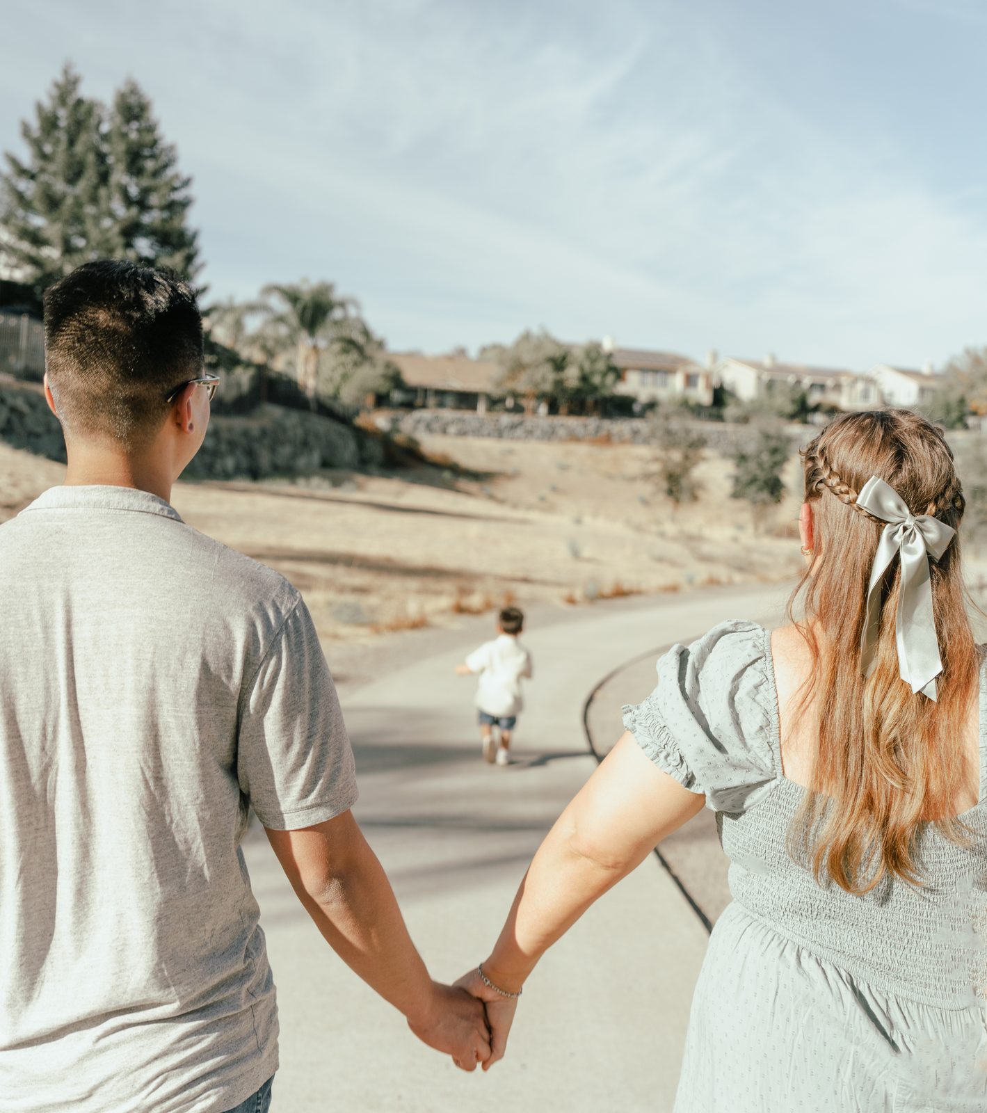 Seen from behind, the parents walk hand in hand down a curving paved path, the mother with a satin bow in her braid, while the toddler is out ahead of them on his own.