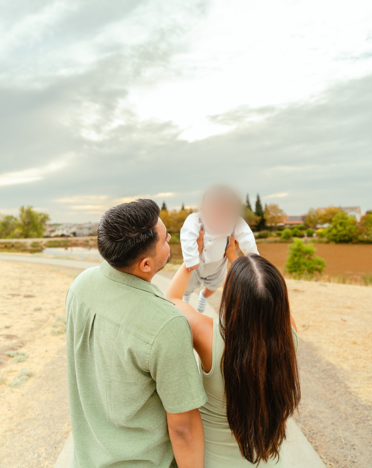 With their backs to the camera, the father and mother stand close on a path as she raises their baby up against a wide, cloudy sky, with rooftops in the distance.
