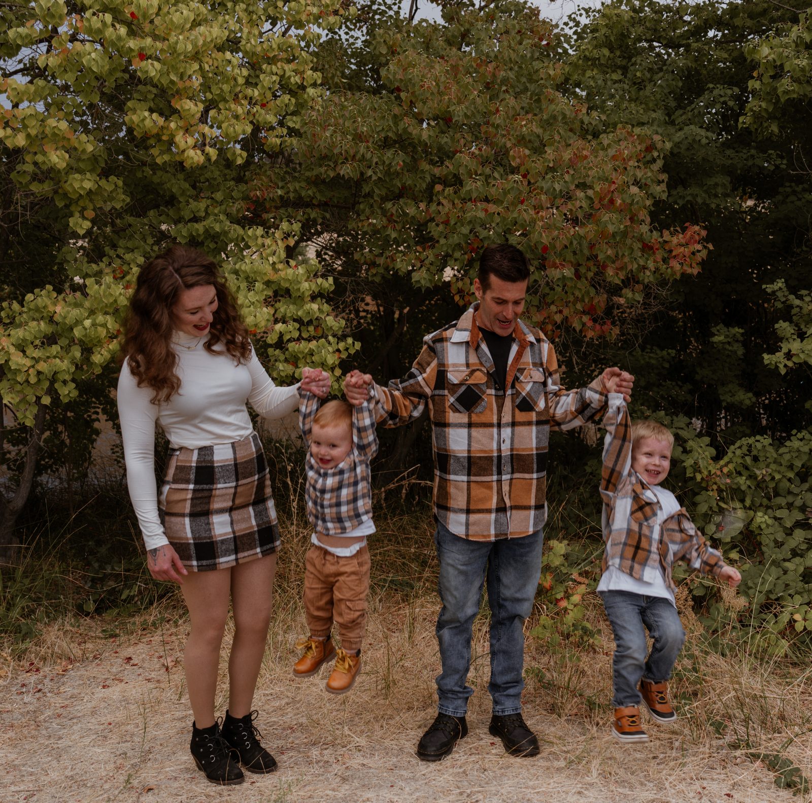 Parents swing their toddler between them by his hands while his older brother hangs from his father's other hand, both boys laughing with their feet off the ground