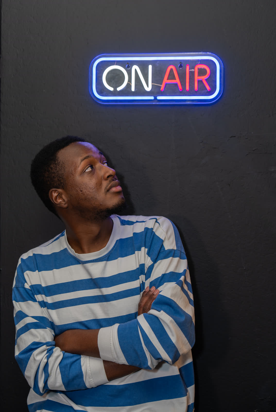 A man in a blue-and-white striped sweater, arms crossed, glancing up at a neon ON AIR sign on a dark wall