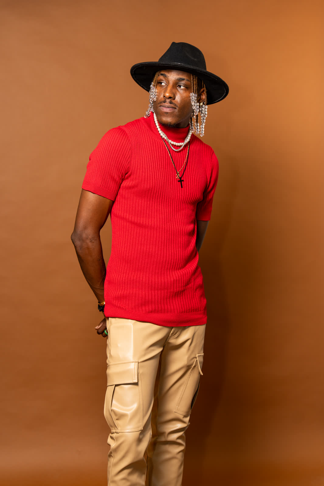 A man in a black wide-brim hat, beaded braids and a red ribbed top, posed against a caramel studio backdrop