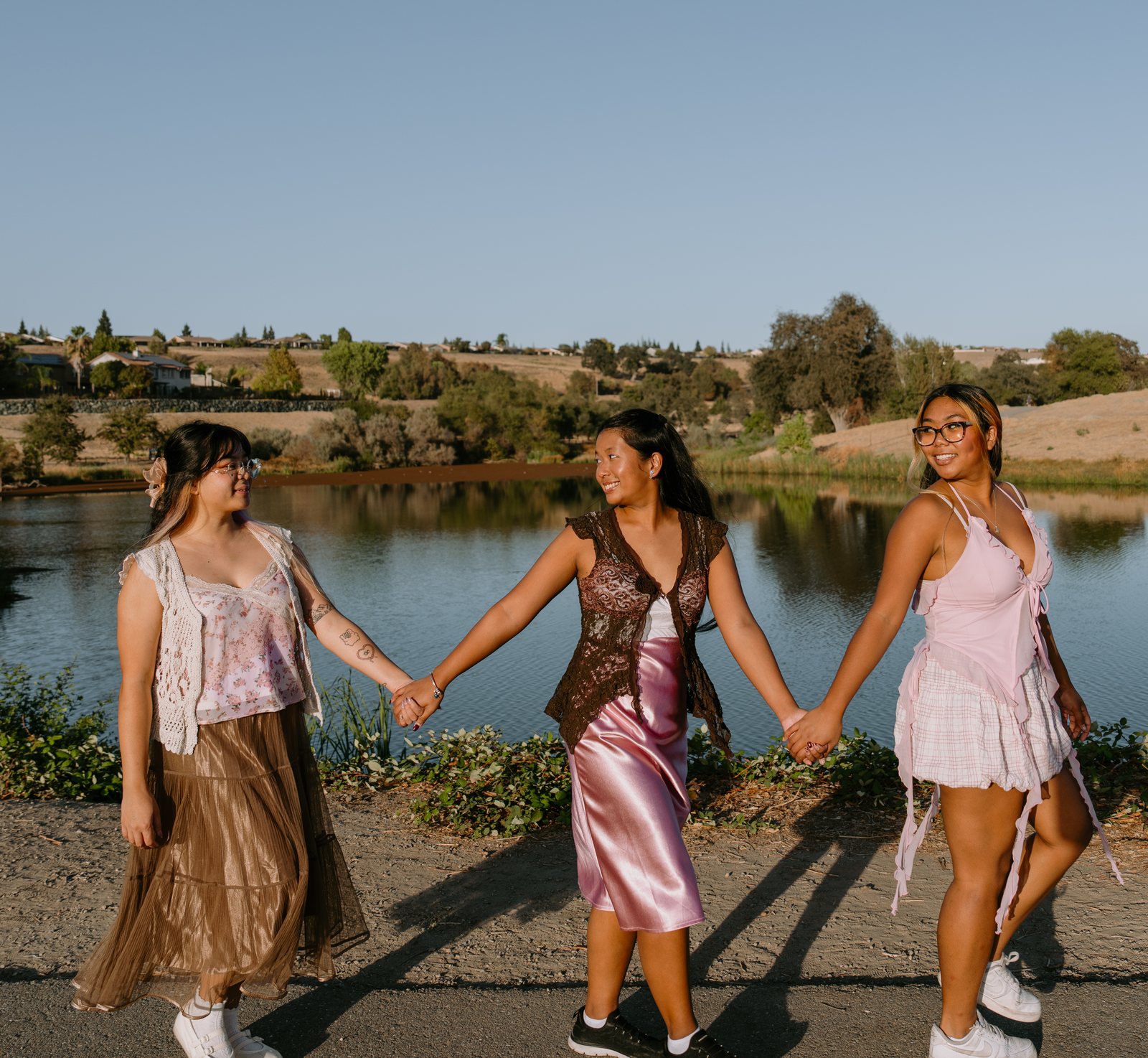 Three young women in pink and brown hold hands in a line on a path beside a pond, two of them smiling at each other, with dry hills and houses across the water.