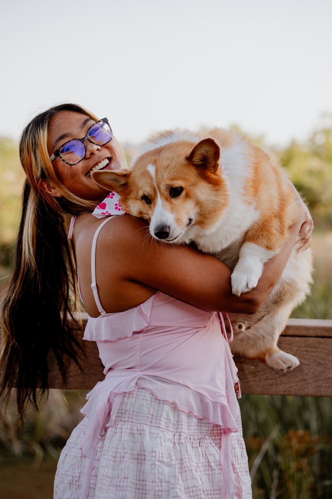A young woman in glasses and a pink top hugs a corgi against her shoulder at a wooden railing and grins back at the camera.